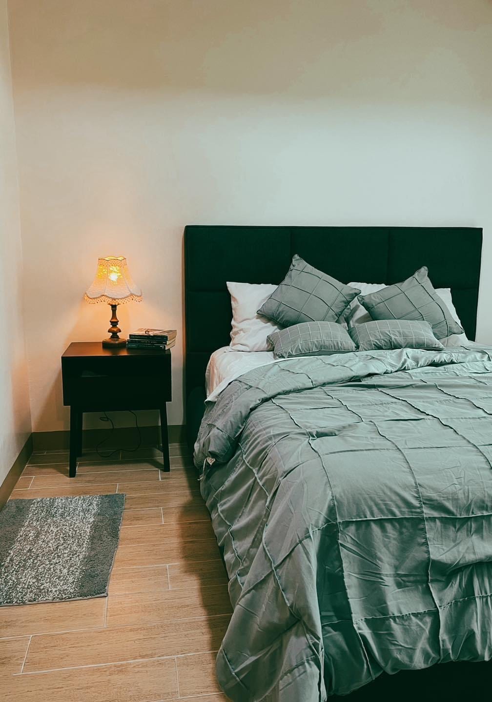 A cozy sleeping area features a dark upholstered headboard with a light gray quilted bedspread. Decorative pillows are arranged on the spacious bed. A bedside table with a vintage lamp provides soft illumination, while a small area rug lies on the floor, enhancing the room's warmth.