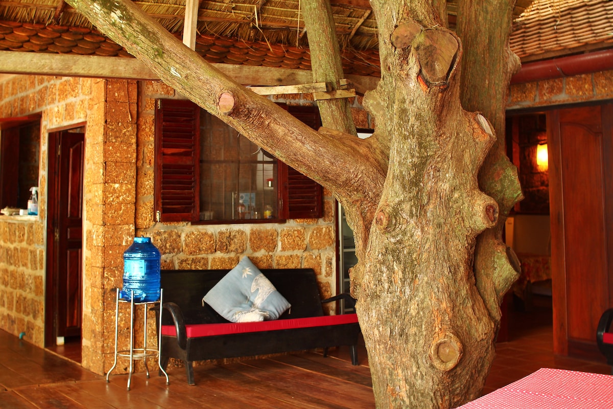 A sturdy tree is positioned prominently in the outdoor seating area, with a black sofa featuring red accents nearby. A blue water dispenser stands on a metallic stand to the side, and natural textures from the stone construction of the villa are visible in the background.