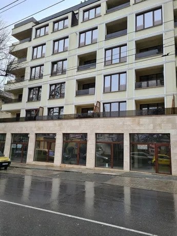 A modern multi-story building is depicted, showcasing a façade of light-colored materials and numerous large windows that provide natural light. The ground floor features expansive glass storefronts. The scene reflects a wet street, suggesting recent rain.