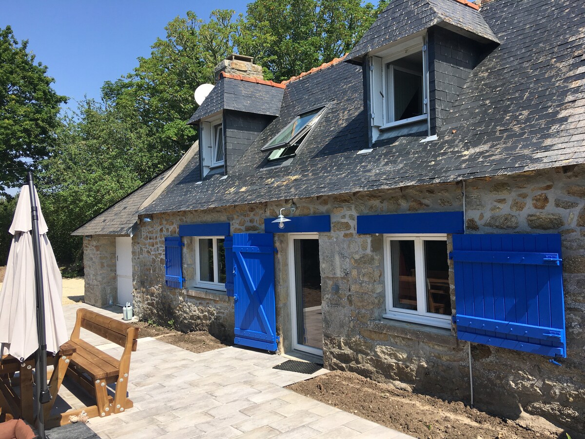 The exterior of the stone cottage is showcased, featuring blue shutters and a dark slate roof. A wooden bench is positioned in front on a stone-paved patio. A parasol stands nearby, providing shade. Lush greenery and trees surround the property, enhancing the peaceful setting.