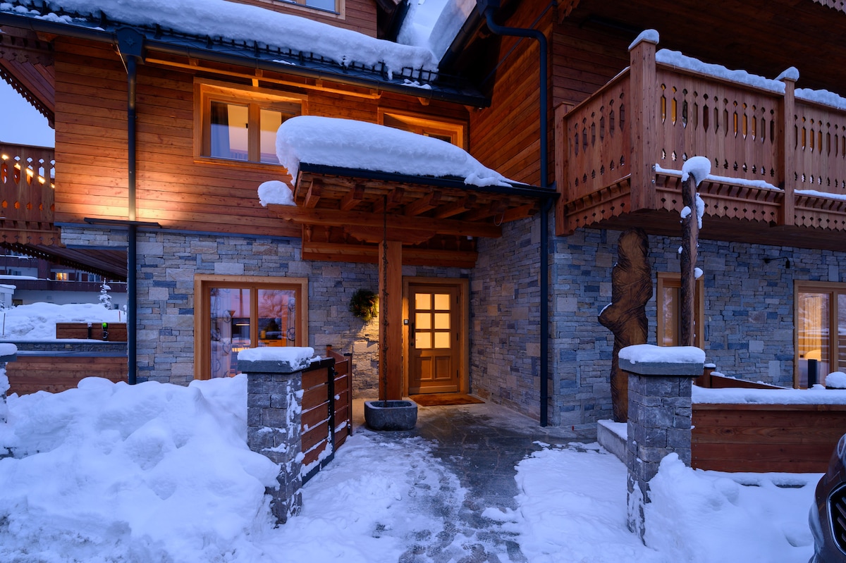 A charming entrance features a wooden door framed by stone walls and snow-covered roof eaves. The path is lined with snow, leading to an inviting entryway illuminated by warm light. Wooden balconies above add character to the rustic design.