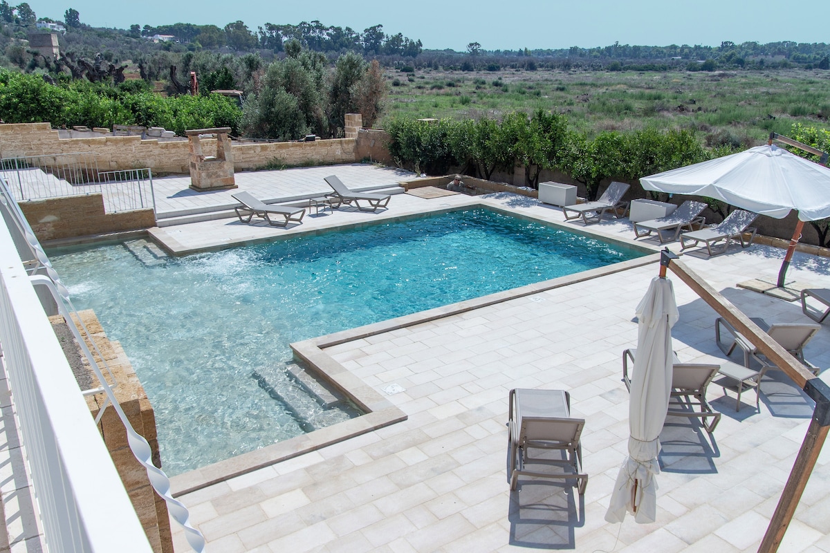 An outdoor pool area features a spacious turquoise pool surrounded by lounge chairs. A large umbrella provides shade nearby, while landscaped greenery and distant trees create a serene backdrop. Sunlight reflects off the water, enhancing the inviting atmosphere.