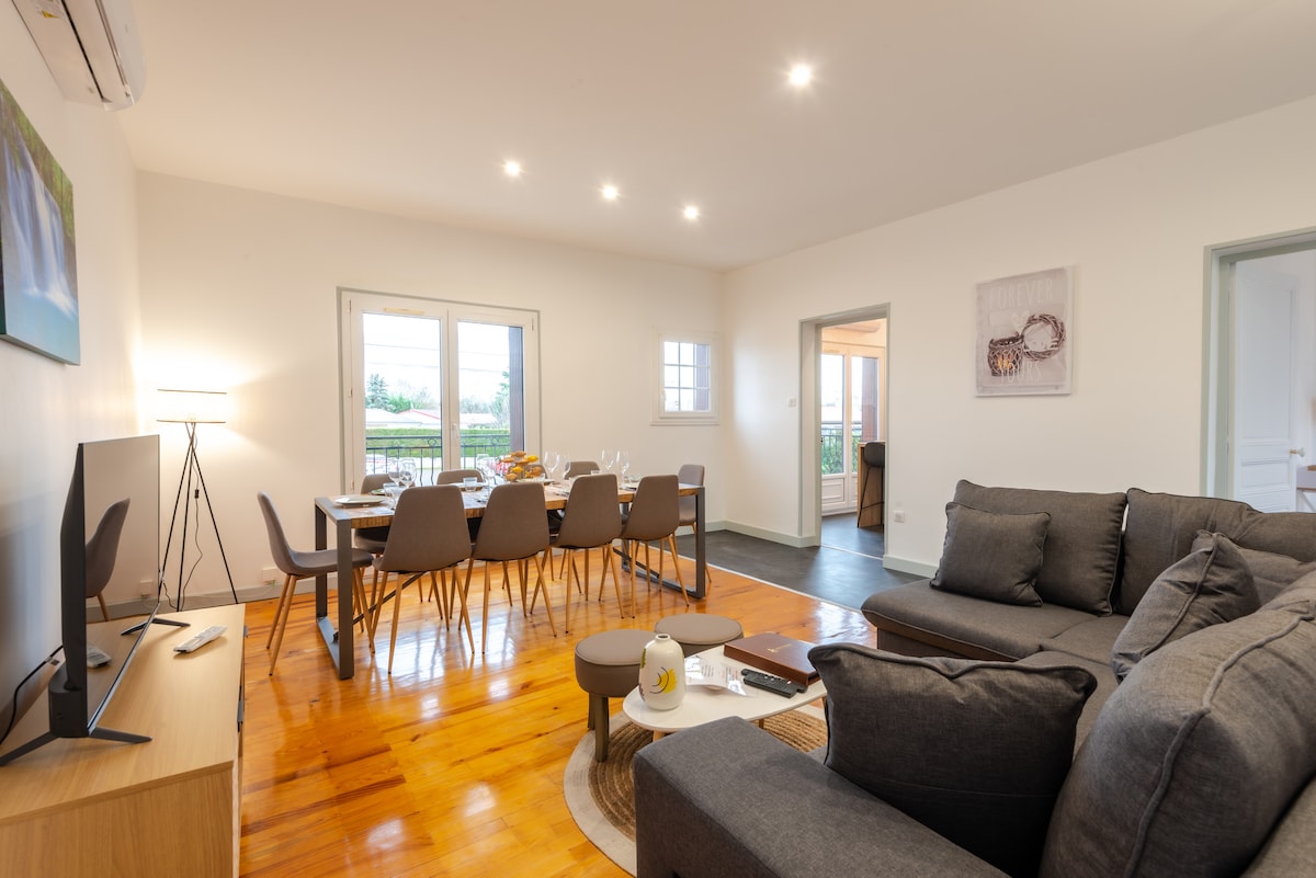 A spacious living area is illuminated by natural light from large windows. A comfortable gray sectional sofa is paired with a side table. A dining table is set for eight, surrounded by modern chairs, with a view of the outdoor landscape.