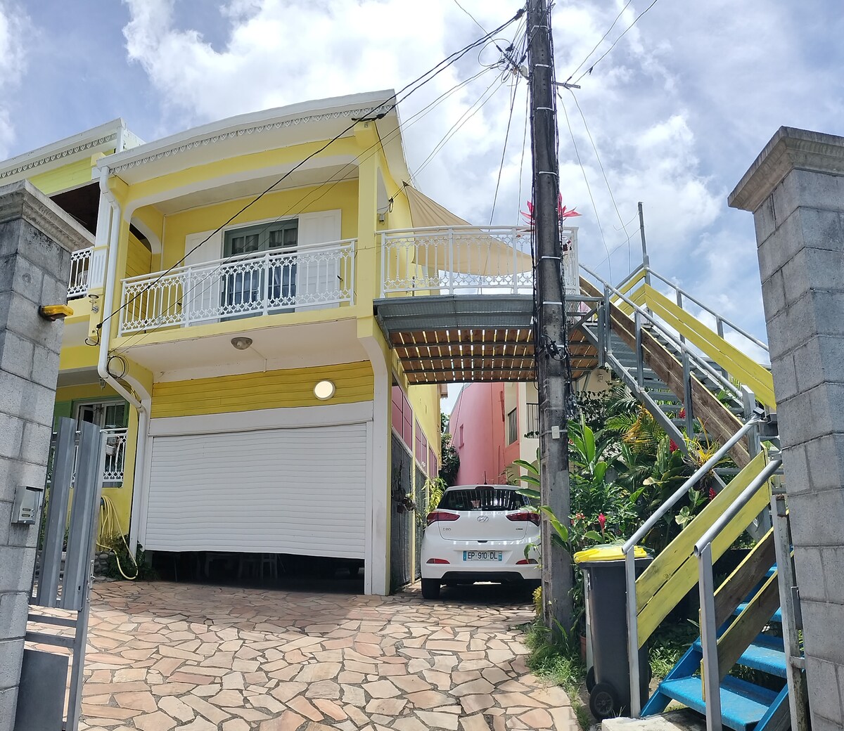 A two-story structure with a bright yellow exterior is accompanied by a stone-paved driveway. A white vehicle is parked in front. An outdoor staircase ascends to the second floor, flanked by greenery, enhancing the welcoming entrance.