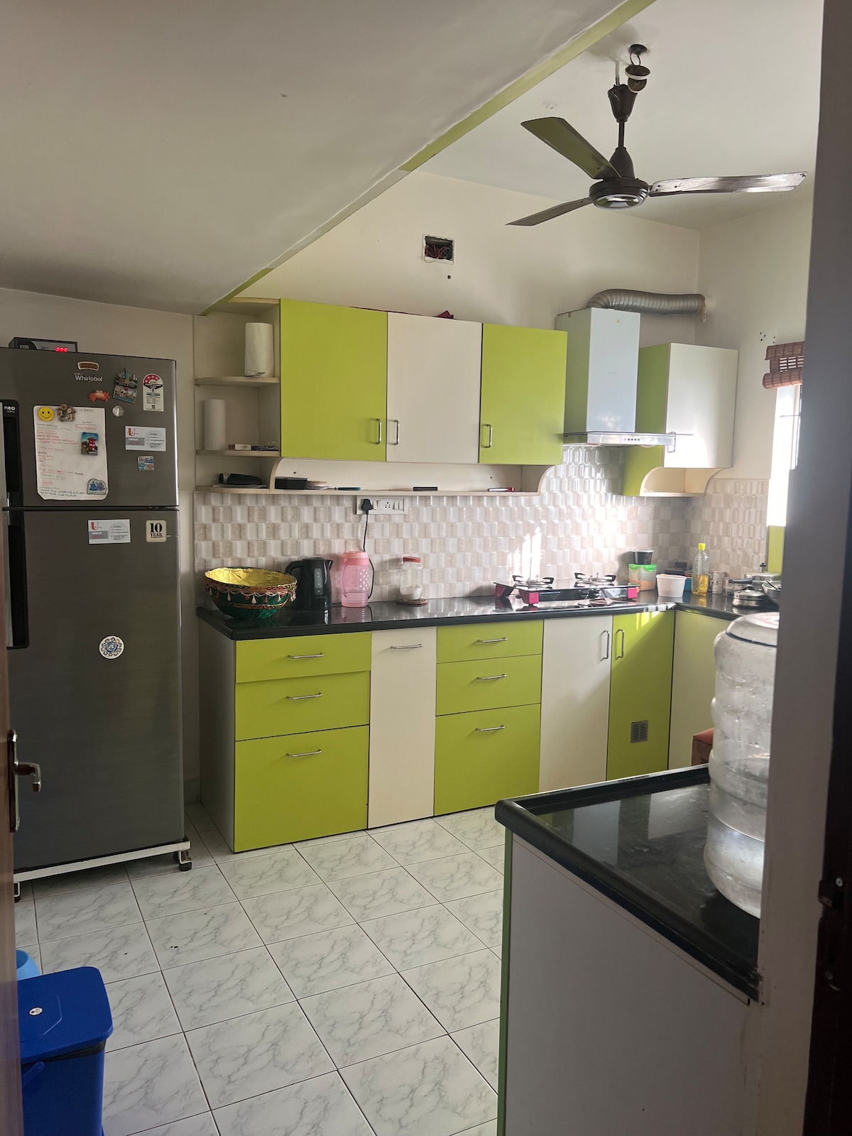 The kitchen features a combination of green and white cabinetry, with ample storage space. Countertops are equipped with essential appliances, including a refrigerator and a gas stove. Natural light filters in through a window, illuminating the clean, tiled floors.