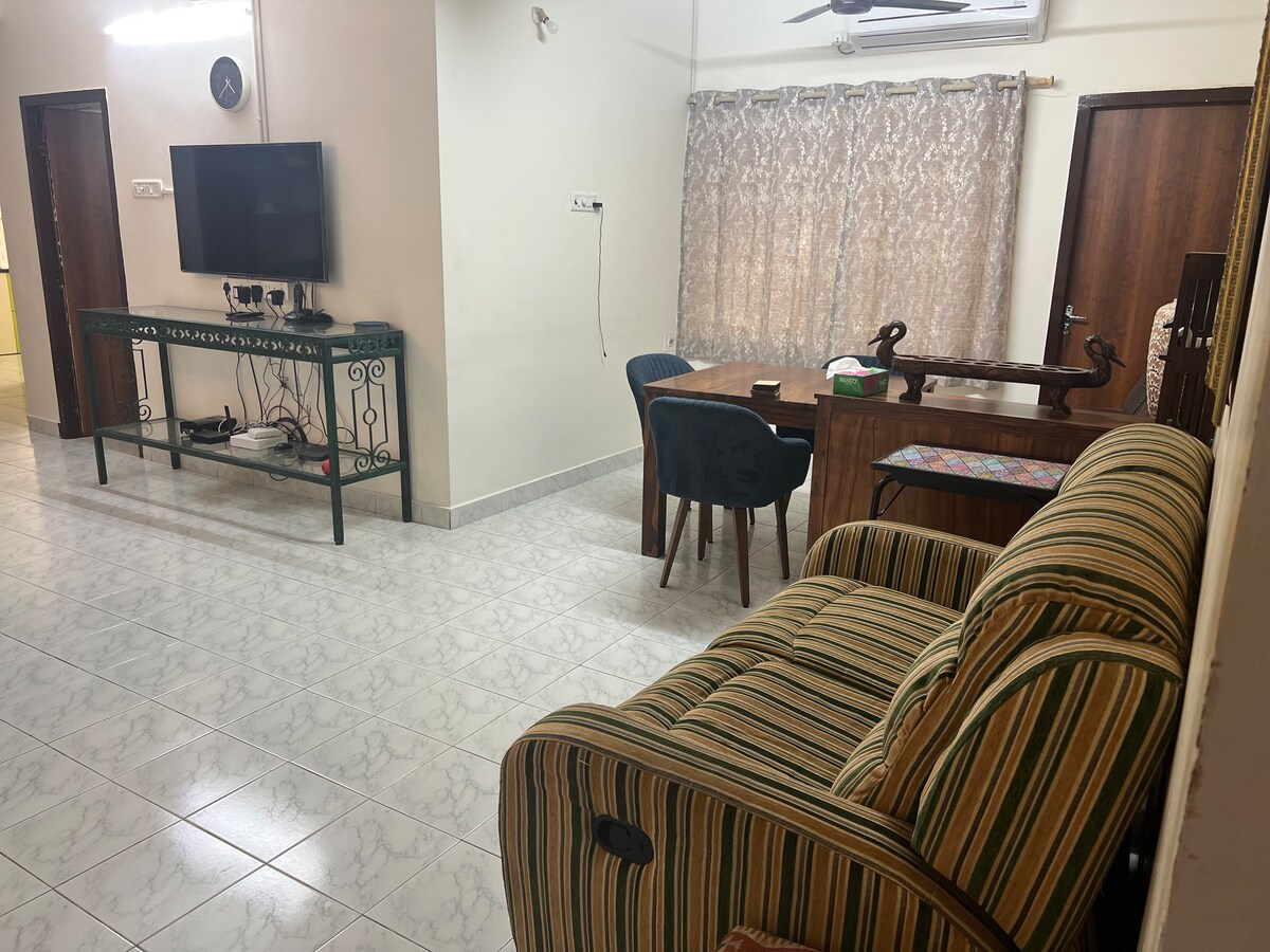A living area features a striped sofa opposite a wall-mounted television. A wooden desk with chair sits next to a window adorned with light, patterned curtains. The floor is tiled, contributing to a spacious and clean ambiance, with warm lighting enhancing the neutral walls.