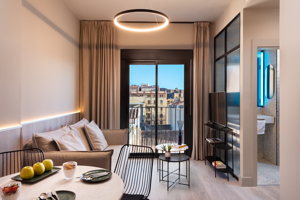 A well-lit living and dining area features a neutral color palette with a comfortable sofa, small round tables, and a dining setup. Large windows offer a view of the city, while modern accents include a contemporary lighting fixture and a flat-screen TV positioned beside the bathroom entrance.