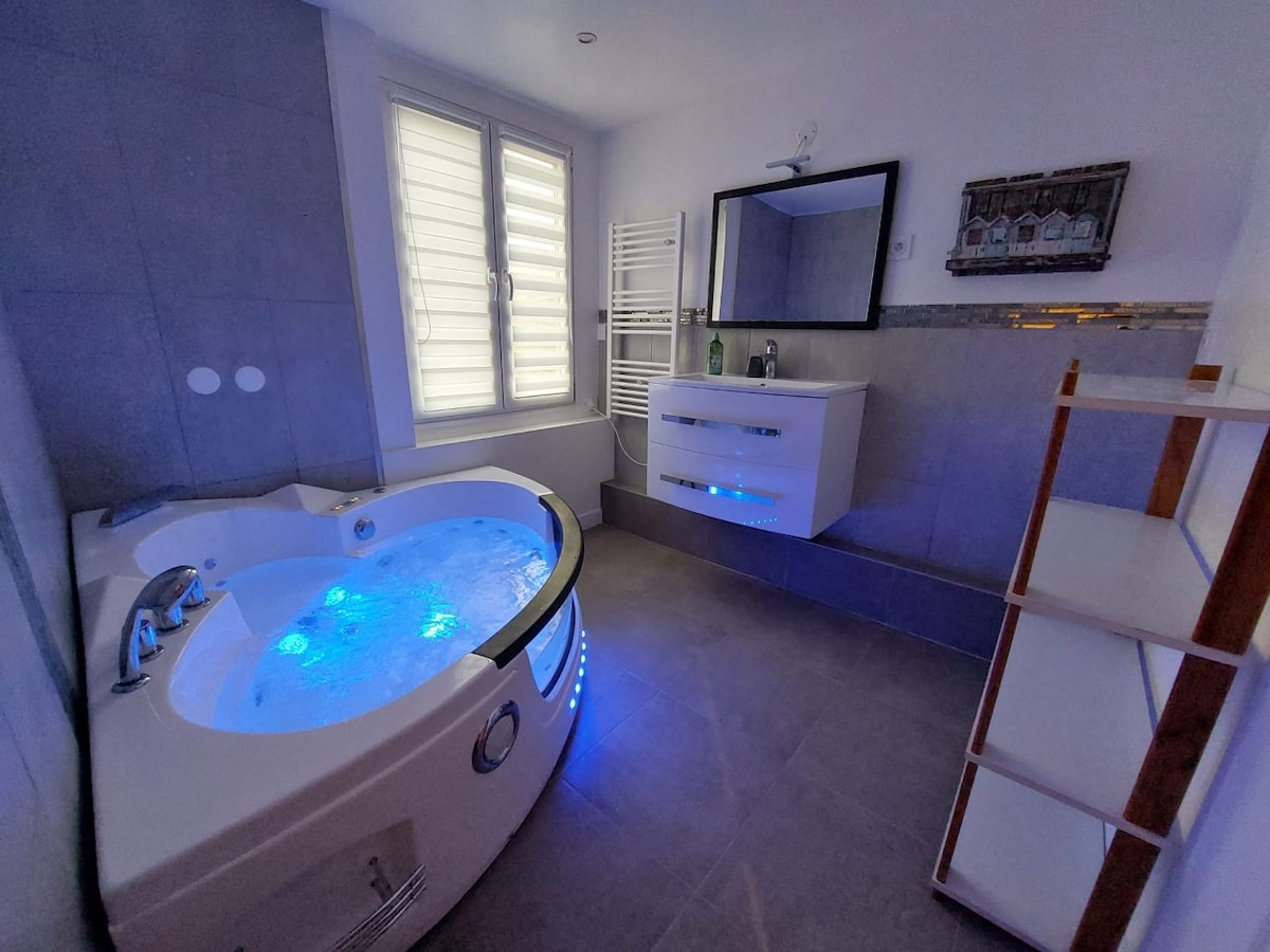 A modern bathroom features a corner whirlpool tub illuminated with soft blue lights. A sleek vanity with a mirror is positioned above a spacious counter, and a towel radiator is mounted against a wall. Natural light enters through a window with white shutters.