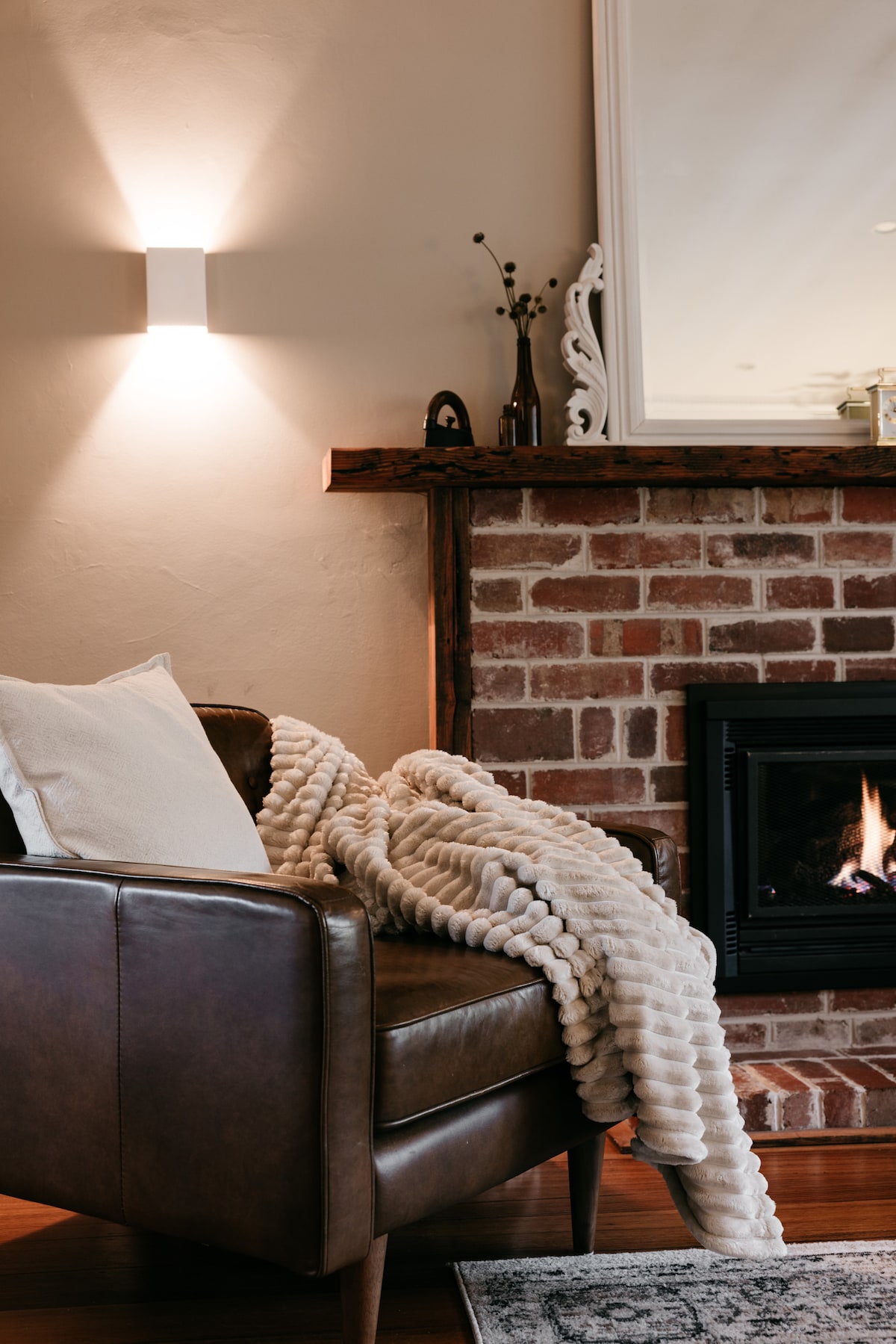 A cozy leather armchair is positioned near a warm gas log fireplace, framed by a rustic brick wall. A soft, textured throw blankets the chair, while a mirror above the mantel reflects the gentle light from a wall-mounted fixture.
