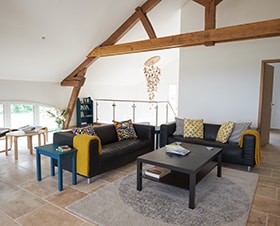 An inviting living area is featured with a low coffee table at the center, surrounded by a dark sectional sofa with colorful cushions. A textured area rug lies beneath, with natural light streaming through large windows adjacent to wooden beams and a stylish chandelier above.