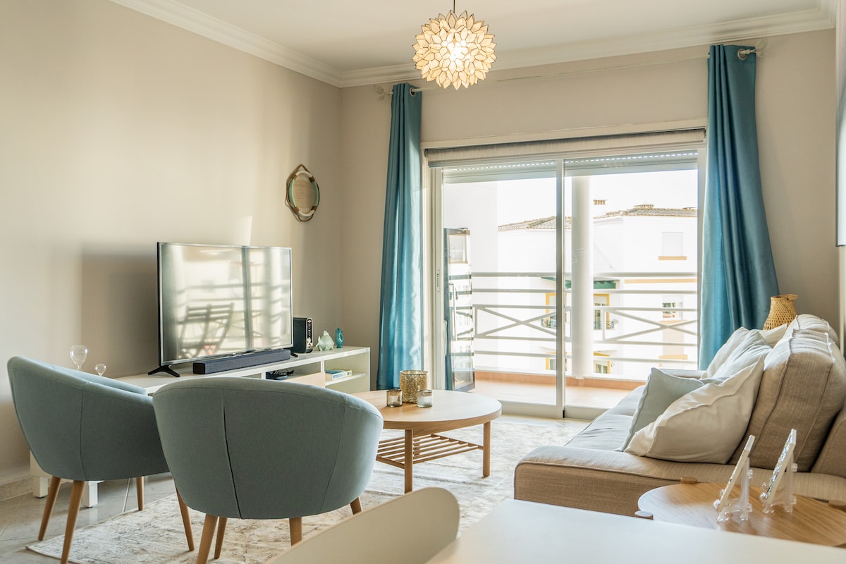 A bright living area is furnished with comfortable seating, including two light blue chairs and a neutral-colored sofa. A sleek television is positioned on a stand, while large sliding doors open to a balcony, allowing natural light to fill the space.
