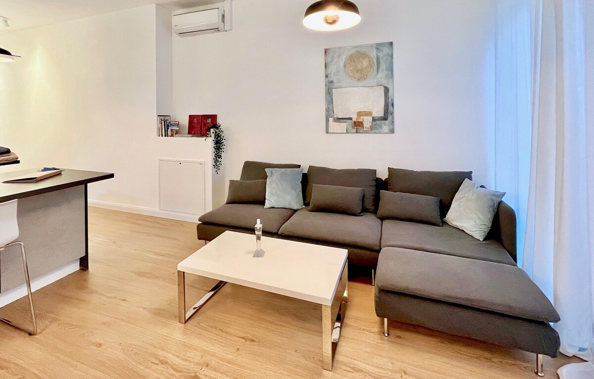 A modern living area features a spacious sectional couch in neutral tones, accented by two decorative pillows. A sleek coffee table sits in front, complemented by a minimalist wall art piece. Natural light floods in through the sheer curtains, enhancing the open layout.