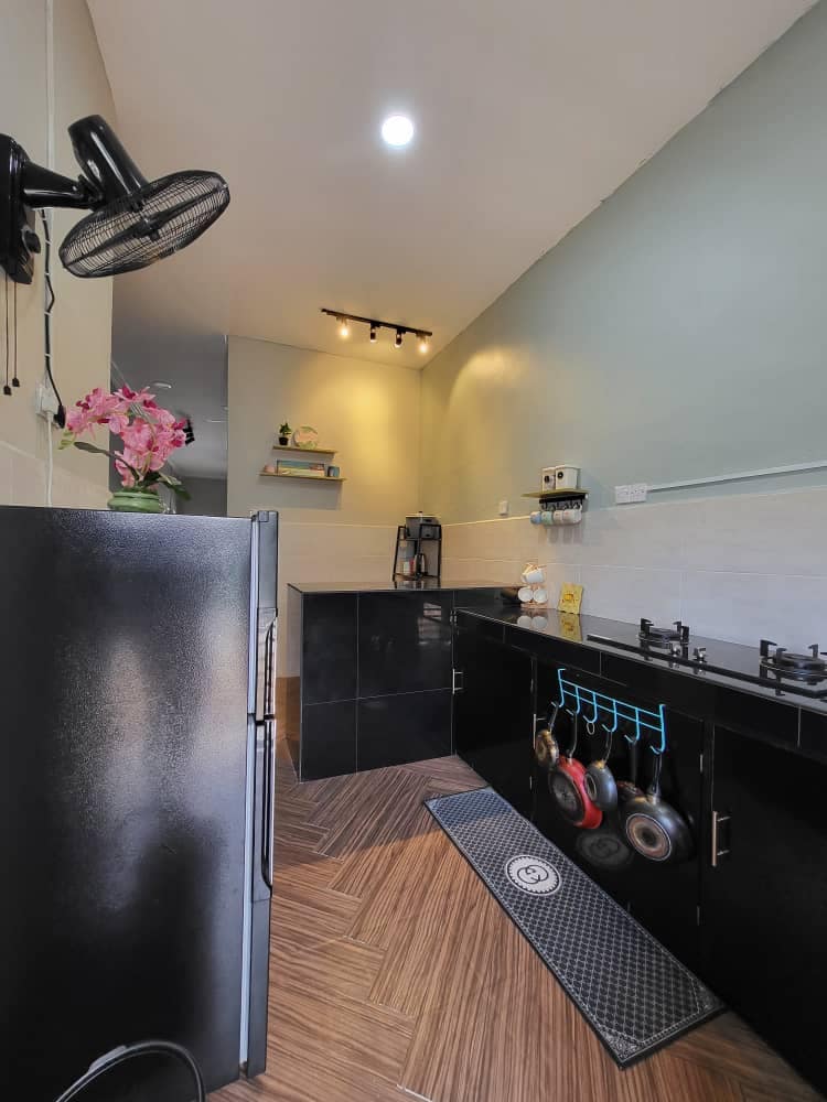 A modern kitchen features sleek black cabinetry with a gas stove and essential cooking utensils displayed. A refrigerator stands on one side, while decorative plants and shelving add a touch of personality, complemented by soft overhead lighting enhancing the functional space.