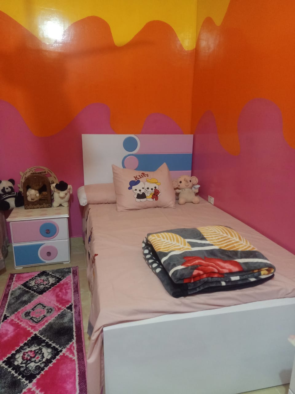 A colorful sleeping area features a single bed adorned with a patterned blanket and a decorative pillow. Bright pink and orange walls create a lively backdrop. A bedside table holds decorative items, while a playful rug is positioned on the floor.