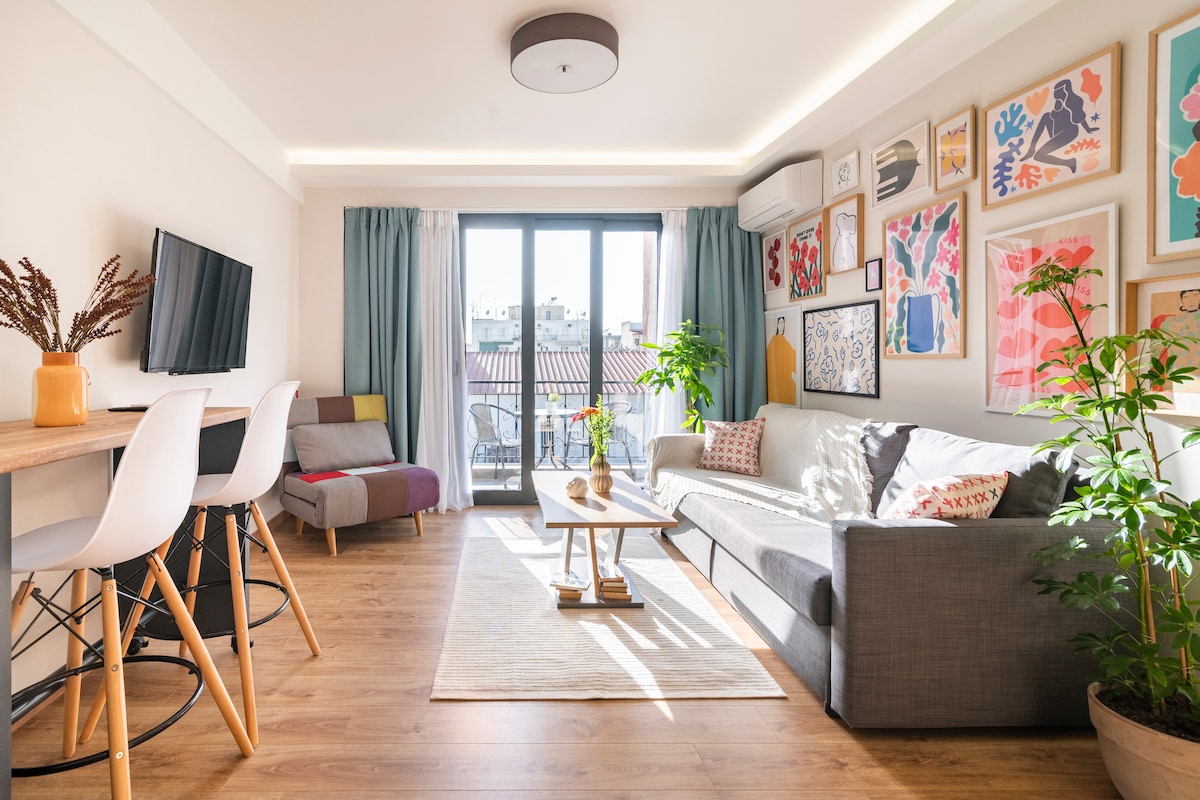 A bright living area features a comfortable gray sofa, a small wooden coffee table, and two white bar stools positioned at a high table. Colorful artwork adorns the walls, while plants are placed in various corners. Large windows allow natural light to fill the space.