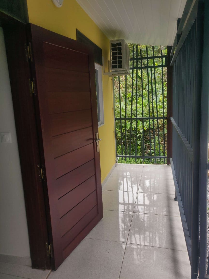A hallway is visible, featuring a modern wooden door with a sleek design. The space is illuminated by natural light reflecting off the polished tile floor. Green foliage can be seen through the open railing, creating a connection to the outdoor surroundings.