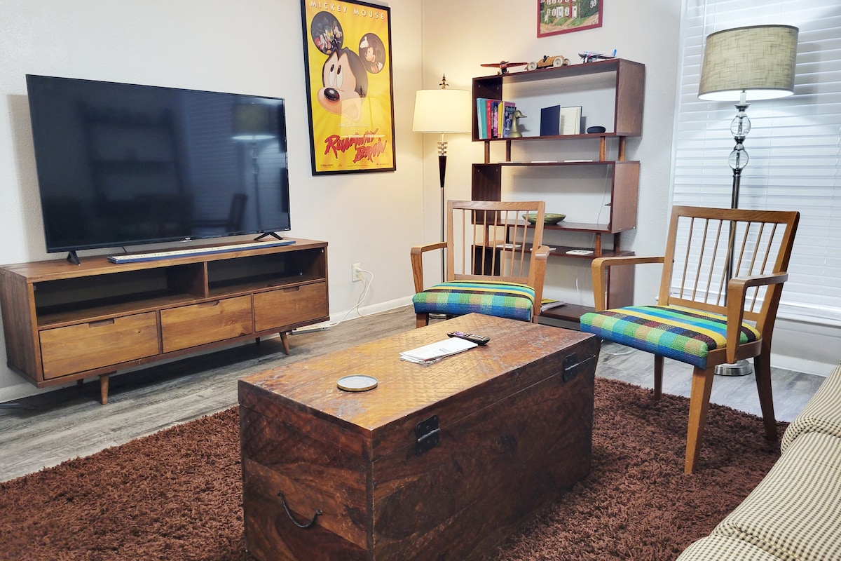 A spacious living area features a 55" 4K Roku TV on a wooden media unit. Two wooden chairs with colorful cushions are positioned beside a rustic coffee table. A soft area rug covers the floor, and a floor lamp provides additional lighting.