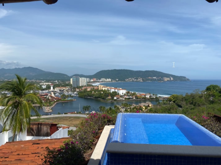 A private terrace features an infinity jacuzzi overlooking a panoramic view of the Pacific Ocean, bay, and mountains. Lush greenery and flowering plants frame the space, while the vibrant coastline and marina are visible in the distance under a clear blue sky.