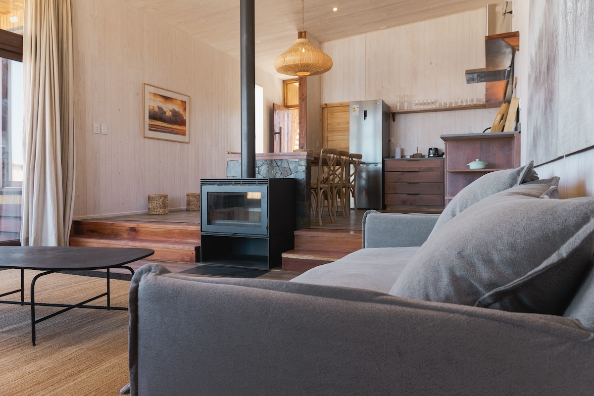 A cozy living area is presented, featuring a modern gray sofa and a low coffee table. A black wood stove stands near the sofa, providing warmth. Natural light enters through large windows, highlighting the open space and contemporary decor.