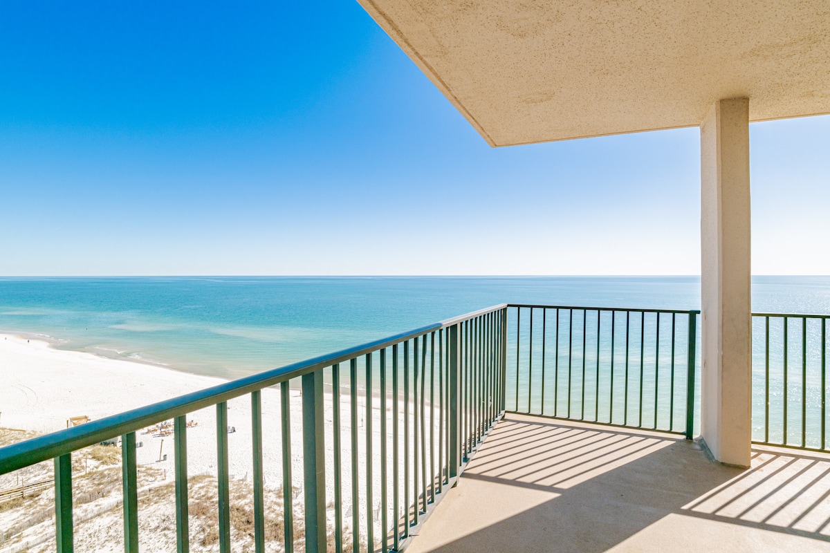 A wrap-around balcony features railings overlooking a serene beach with soft white sand. The calm blue waters extend to the horizon under a clear sky, allowing for unobstructed views of the tranquil seaside environment.