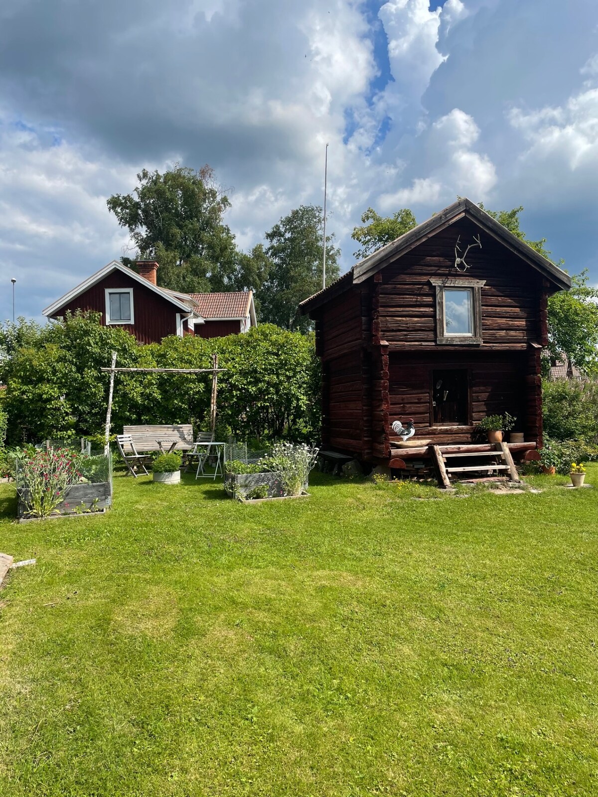 A charming wooden cabin is framed by lush greenery in a well-maintained garden. The structure features a small staircase leading to the entrance, with a decorative window visible on the front. In the background, a larger house can be seen, surrounded by foliage.