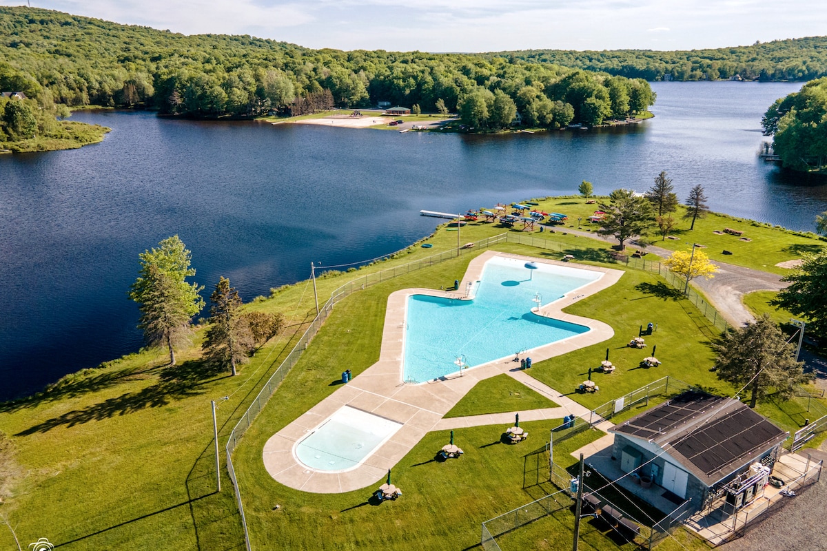 An aerial view showcases a large, outdoor swimming pool surrounded by expansive green lawns. The calm lake reflects the lush tree line, enhancing the serene environment. Nearby, recreational equipment is visible along the shores, contributing to the community's outdoor activities.