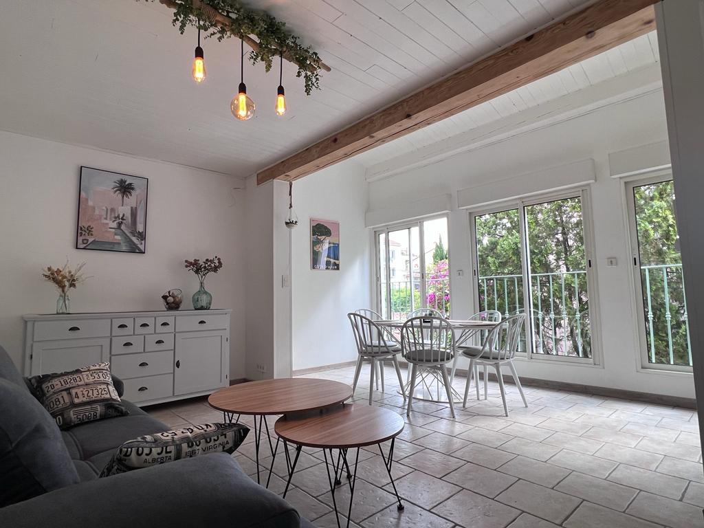 A living space is presented with large windows allowing natural light to fill the room. A comfortable seating area features a gray sofa. Two round wooden tables are displayed next to a dining area, furnished with a round table and white chairs. Decorative plants are positioned throughout.