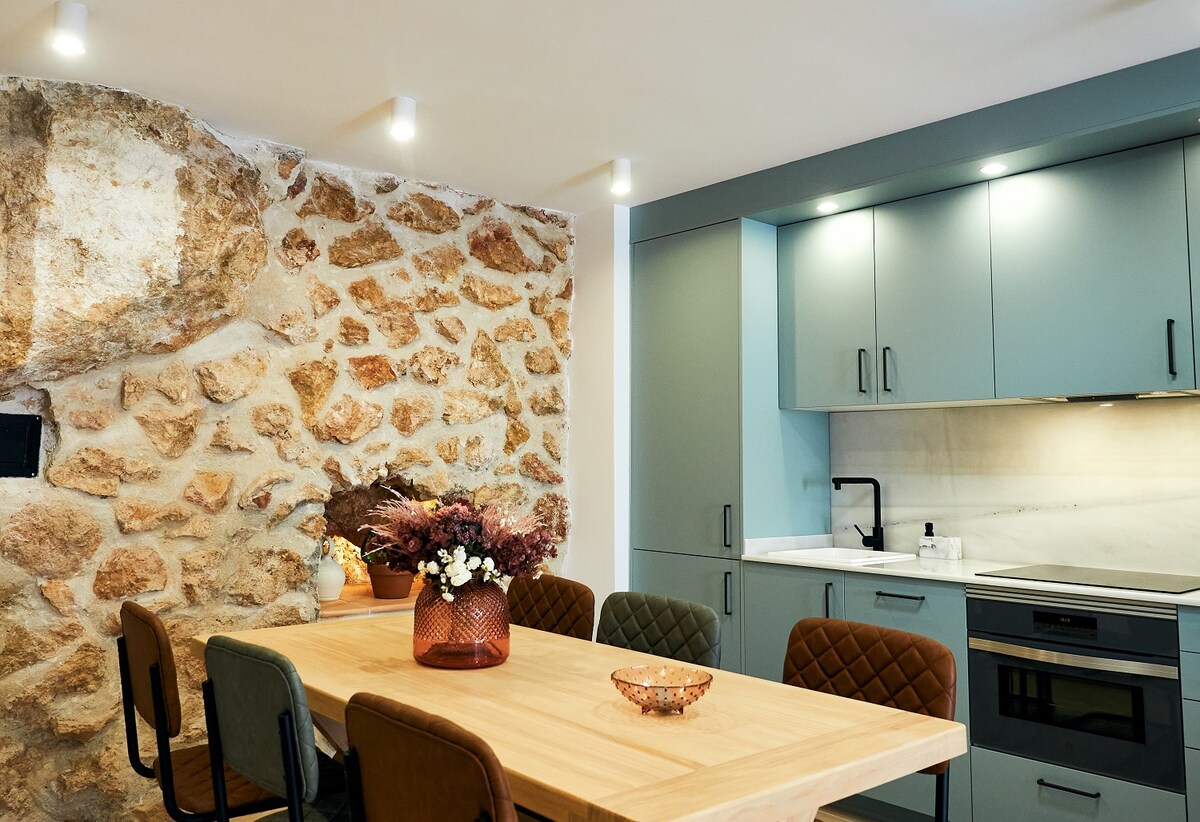 A modern kitchen is showcased with a combination of stone and sleek cabinetry. A wooden dining table surrounded by variously colored chairs invites gathering. The kitchen features contemporary appliances and soft lighting, enhancing the functional space.