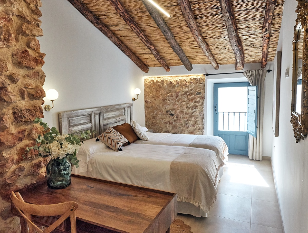 The sleeping area features two spacious beds with neutral linens, complemented by decorative pillows. Exposed wooden beams and stone walls add a rustic charm. A wooden desk is positioned nearby, and a window offers natural light with views of the outside.