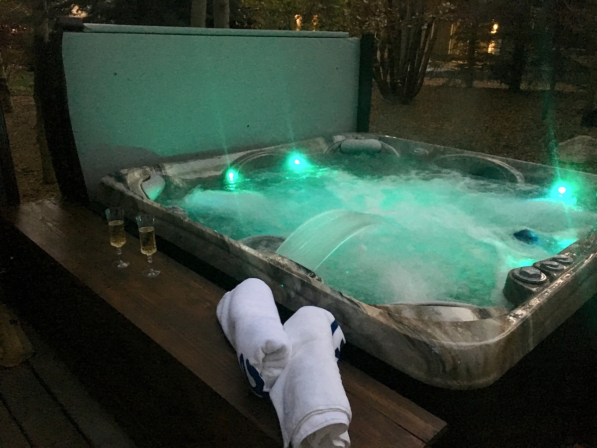 An outdoor hot tub is positioned on a wooden deck, surrounded by trees. Soft green lighting casts a serene glow on the bubbling water. Two glasses are placed nearby, alongside neatly folded towels, contributing to a relaxing ambiance.