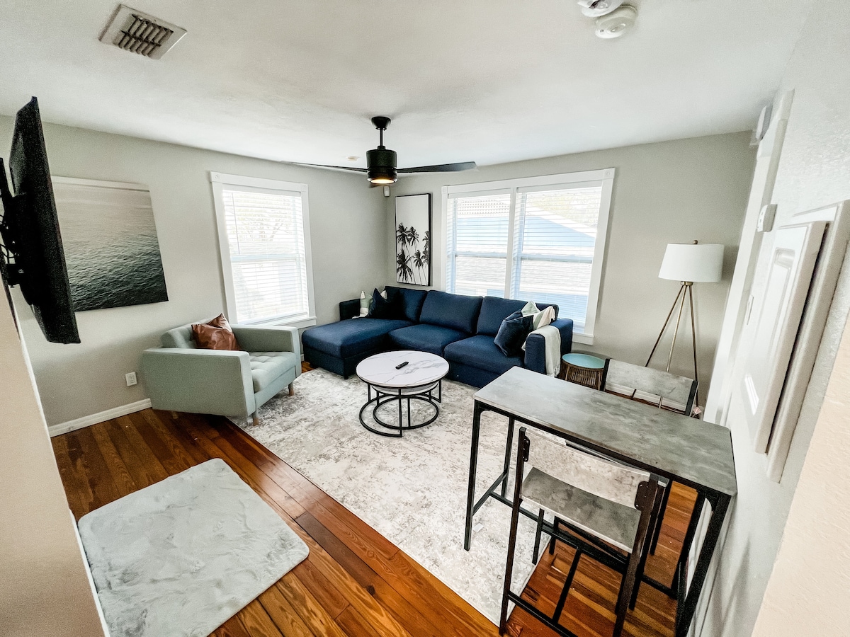 An open living area is shown, featuring a large blue sectional sofa, a round coffee table, and a small dining table with two chairs. Light enters through three windows, illuminating the hardwood floor and neutral walls. An accent chair and a floor lamp provide additional seating and lighting.