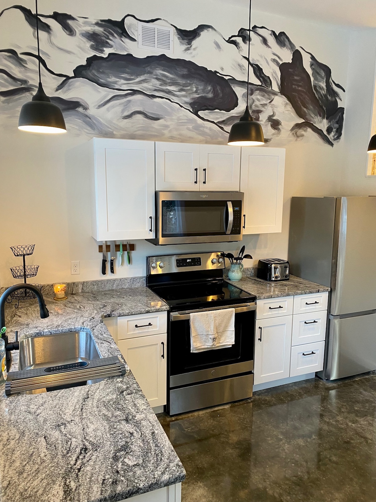 The kitchen features a stylish design with white cabinetry and modern stainless steel appliances, including a microwave and refrigerator. A hand-painted mountain mural adorns the wall above a granite countertop, complemented by black pendant lighting and a functional sink.