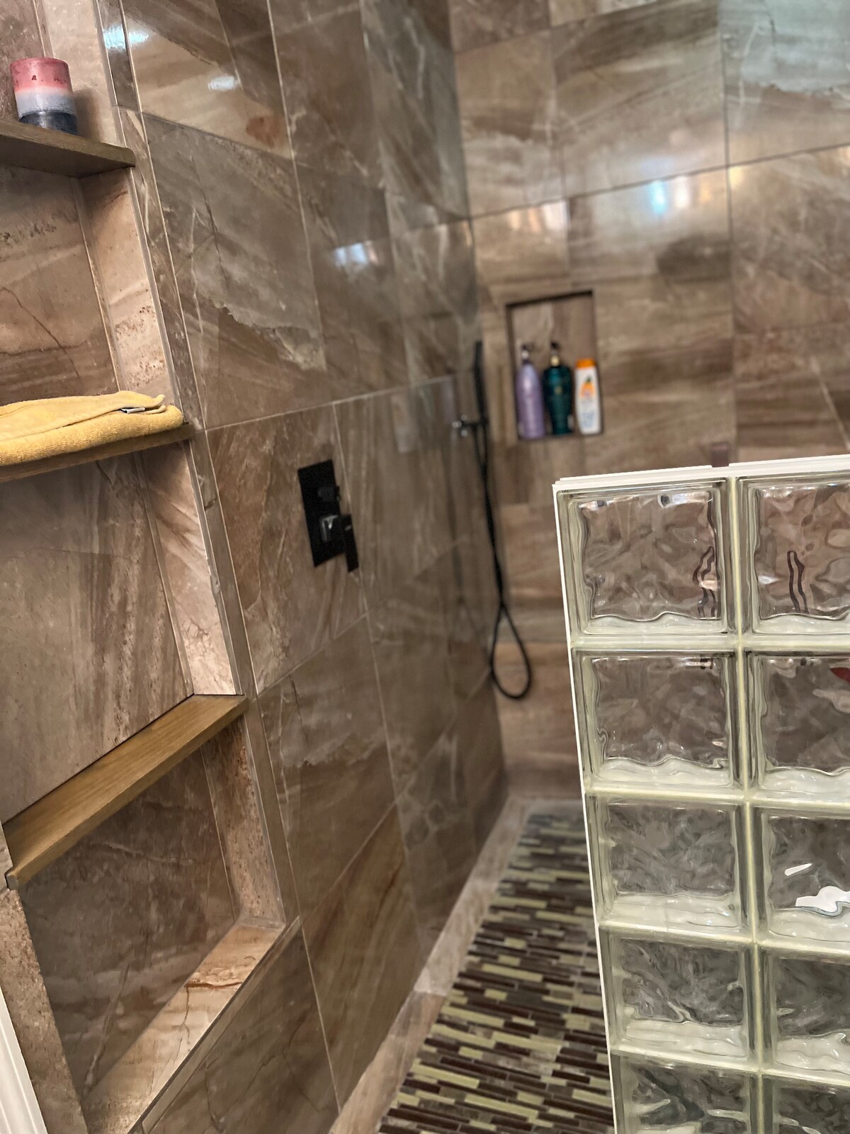A modern shower area features textured brown tiles and built-in shelves holding toiletries. A clear glass block wall enhances the space, allowing light while maintaining privacy. A patterned mat lies on the floor, contributing a touch of warmth to the sleek design.