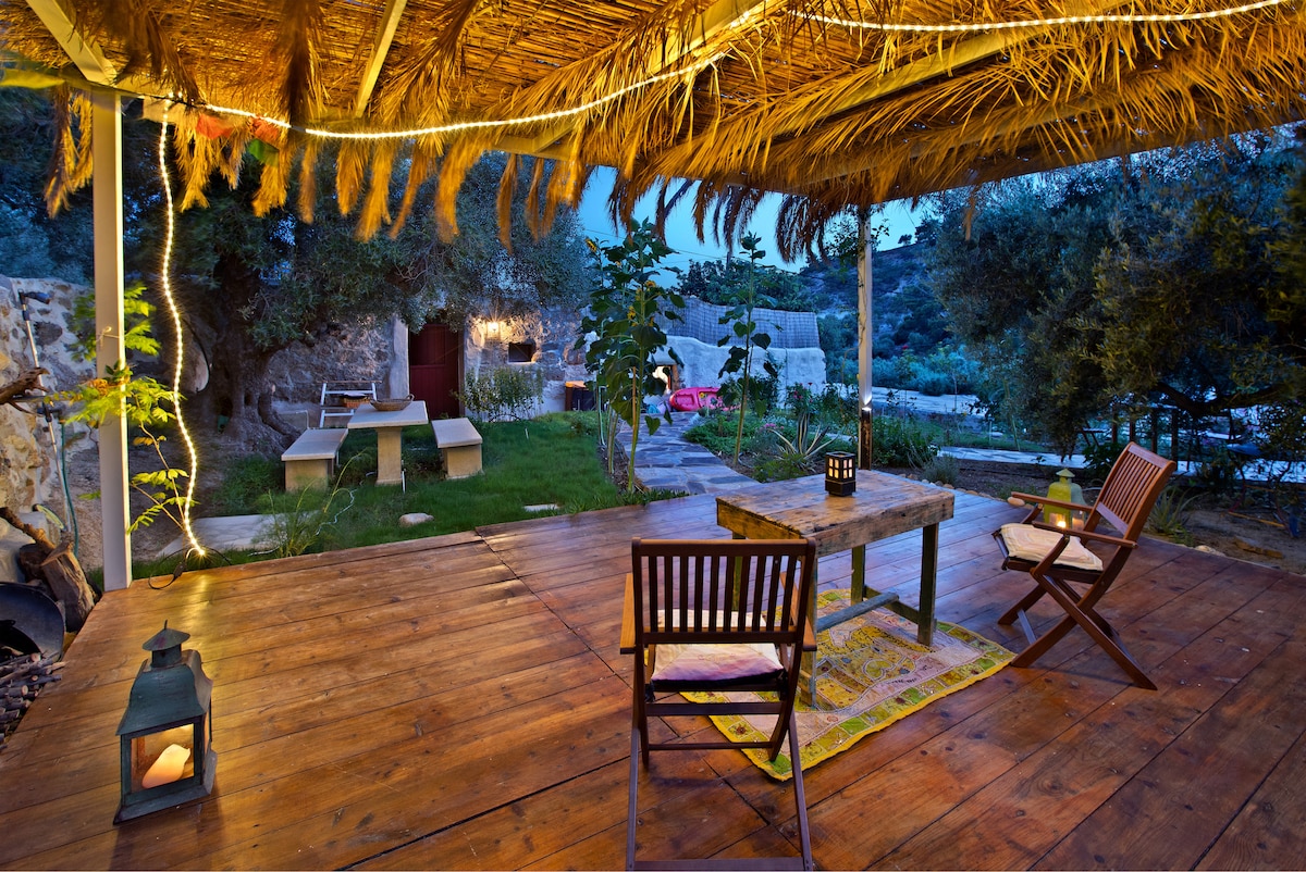 An outdoor veranda is covered by a thatched roof, featuring wooden flooring and illuminated by soft lighting. A wooden table and two folding chairs are arranged in the center, while lush greenery and stone pathways are visible in the background, enhancing the serene atmosphere.