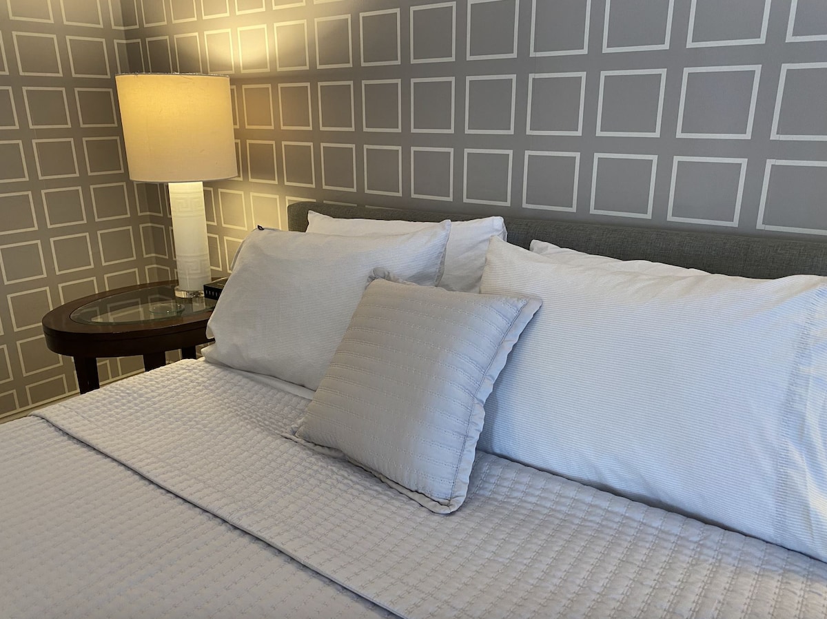 A close-up view of a neatly arranged bed featuring white bedding and an accent pillow. A bedside table with a lamp is visible, providing soft illumination, while a patterned gray wall adds texture to the cozy sleeping area.