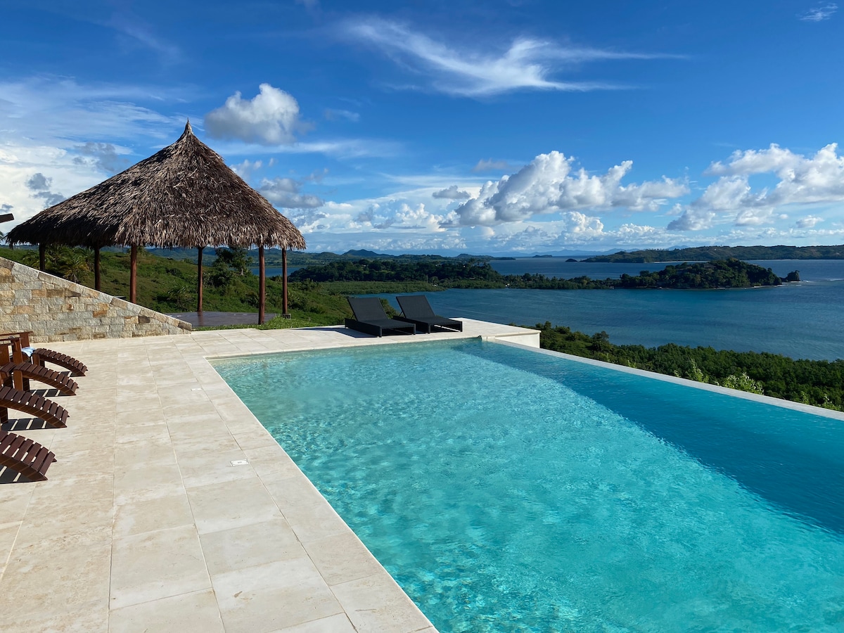 An infinity pool is surrounded by a stone terrace, offering views of the landscape and water. A thatched gazebo provides shaded seating. Two lounge chairs are placed beside the pool, integrating seamlessly with the tranquil environment.