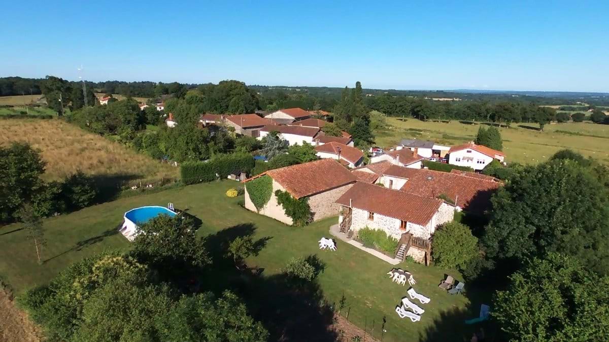 An aerial view captures the tranquil landscape featuring multiple stone gîtes surrounded by green gardens. A shared heated pool is visible alongside lounge chairs and a well-maintained lawn, with fields stretching into the distance under a clear blue sky.
