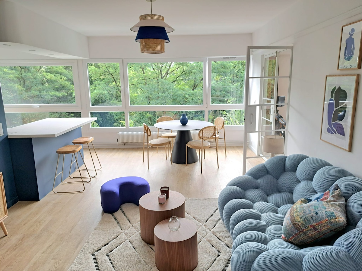 A bright living area features large windows offering views of surrounding greenery. A cozy blue sofa is complemented by two wooden tables. A dining table with chairs and two barstools are arranged near the kitchen counter, enhancing the inviting space.