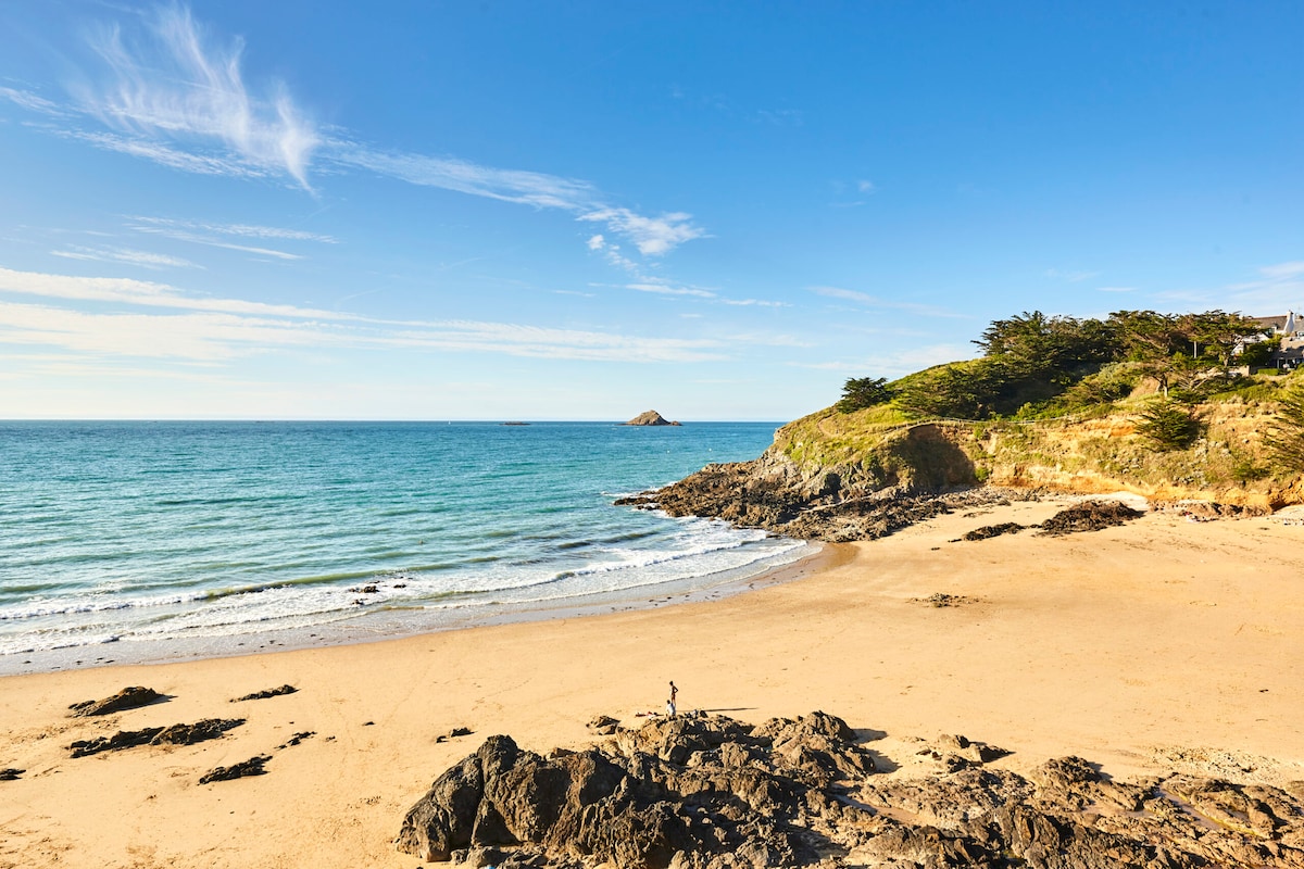 A serene beach scene is depicted, showcasing soft sandy shores and gentle waves lapping against the coastline. Rock formations are visible, and a distant island can be seen in the open water under a clear blue sky with light clouds.
