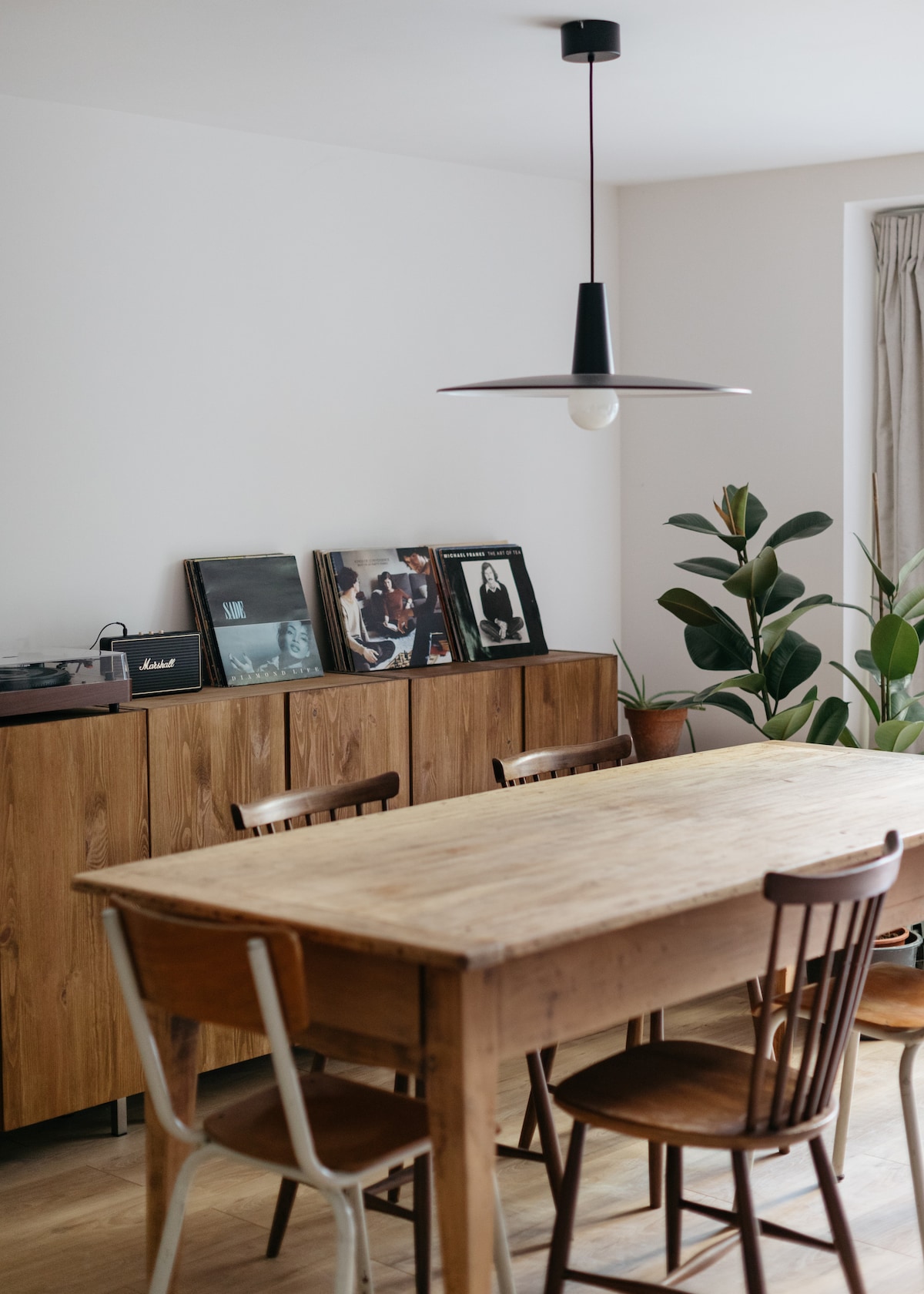 A wooden dining table with several chairs is positioned in the center of the room. A record player and vintage vinyl records are displayed on a cabinet against the wall. Potted plants add a touch of greenery, complementing the natural wood tones.