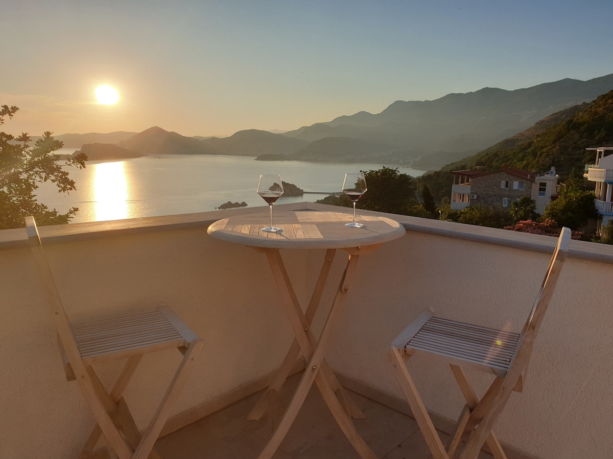 A small balcony features a round wooden table and two matching chairs, positioned to enjoy a stunning sunset view. The sun casts a warm glow over the water and mountains in the distance, creating a serene setting for relaxation.