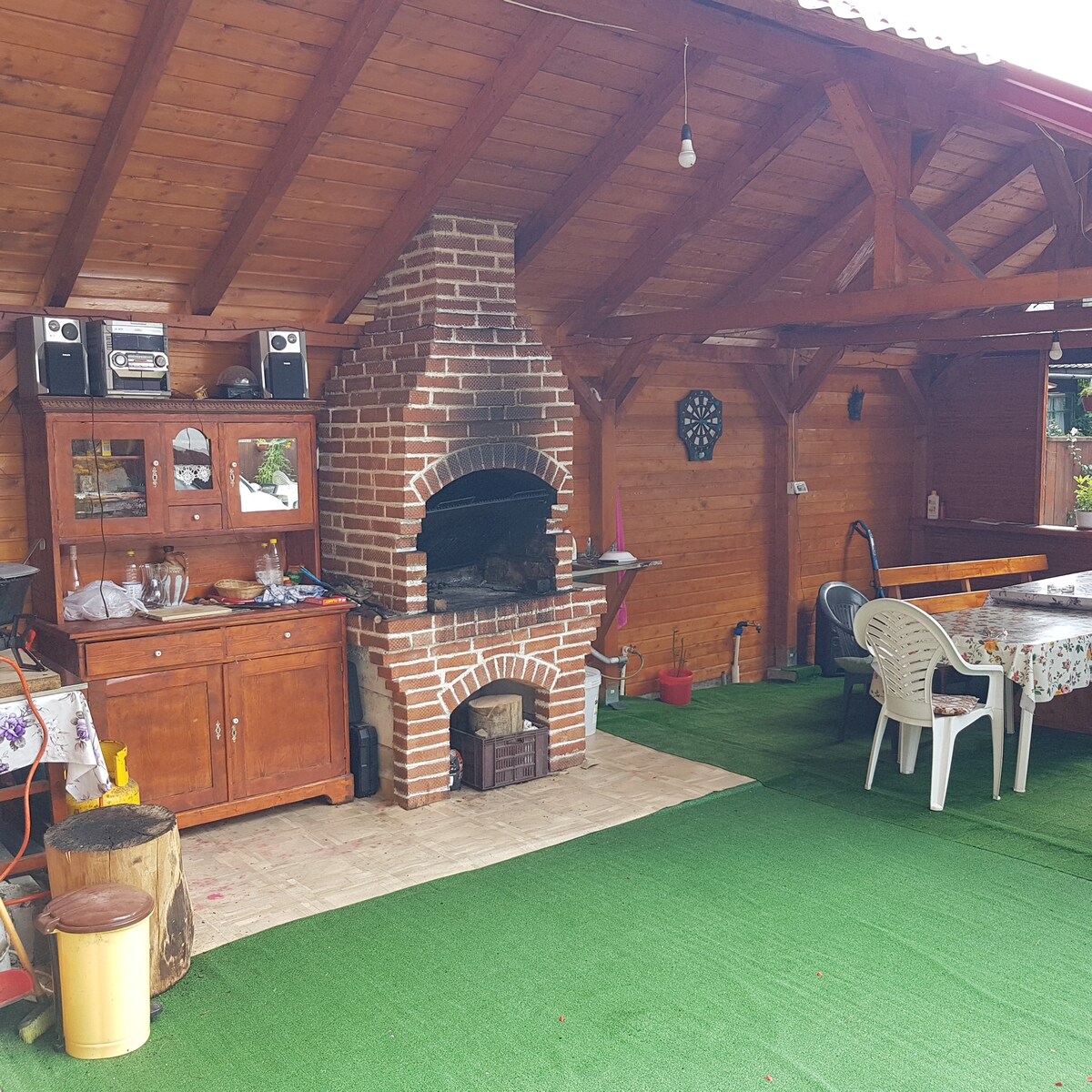 An outdoor covered space features a brick barbecue grill as a central focal point. Wooden cabinets and a table with chairs line the area, while a green carpet adds comfort underfoot. Natural wood beams and an open roof create an inviting atmosphere for gatherings.