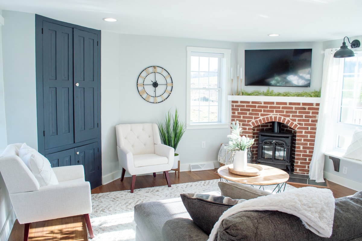 A cozy living area is highlighted by a gas stove fireplace set within a charming brick wall. Light green walls complement a soft area rug and a round wooden coffee table. Two comfortable chairs are positioned invitingly, and natural light flows through large windows.