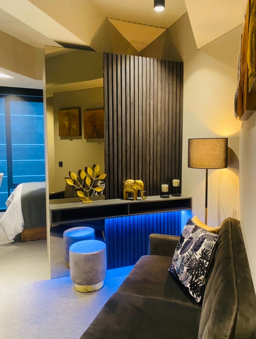 The interior space features a cozy seating area with a dark sofa adorned with decorative cushions. A wall with vertical wooden slats adds texture and interest. A sleek lamp provides soft lighting, while a console table displays decorative items, enhancing the modern aesthetic of the studio.