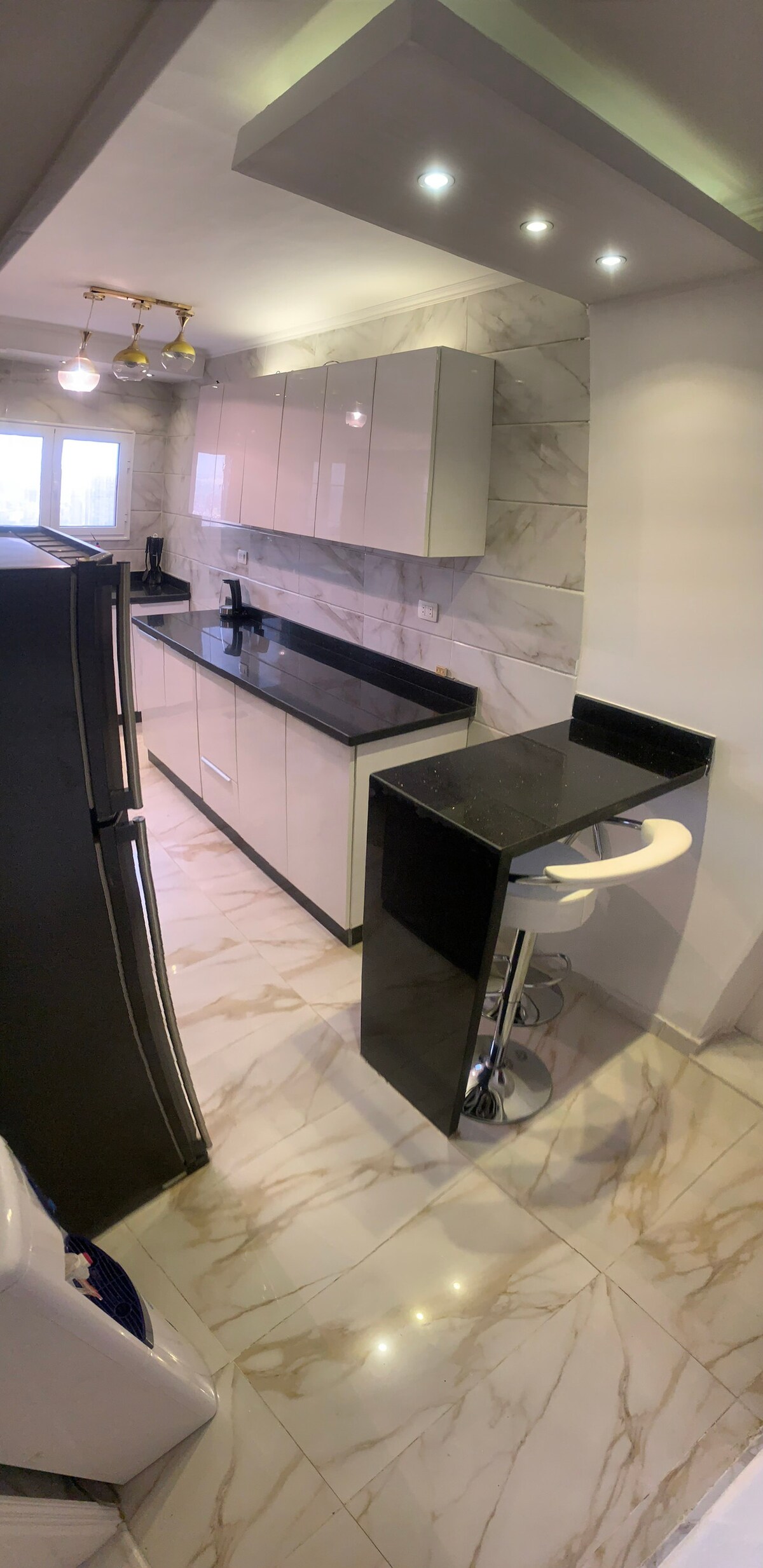 A modern kitchen features sleek white cabinets paired with a black countertop. A compact bar area with a stool is positioned near a window, allowing natural light to brighten the space. Glossy tiles reflect the kitchen's contemporary design.