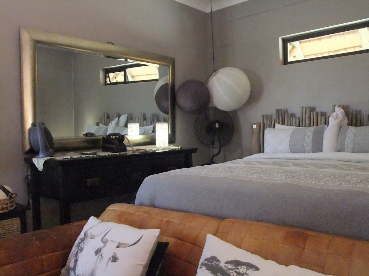 A spacious sleeping area features a large bed dressed in soft linens and adorned with decorative pillows. A mirrored dresser reflects light and adds depth. Unique paper lanterns hang overhead, and a comfortable seating area with textured cushions is positioned nearby.
