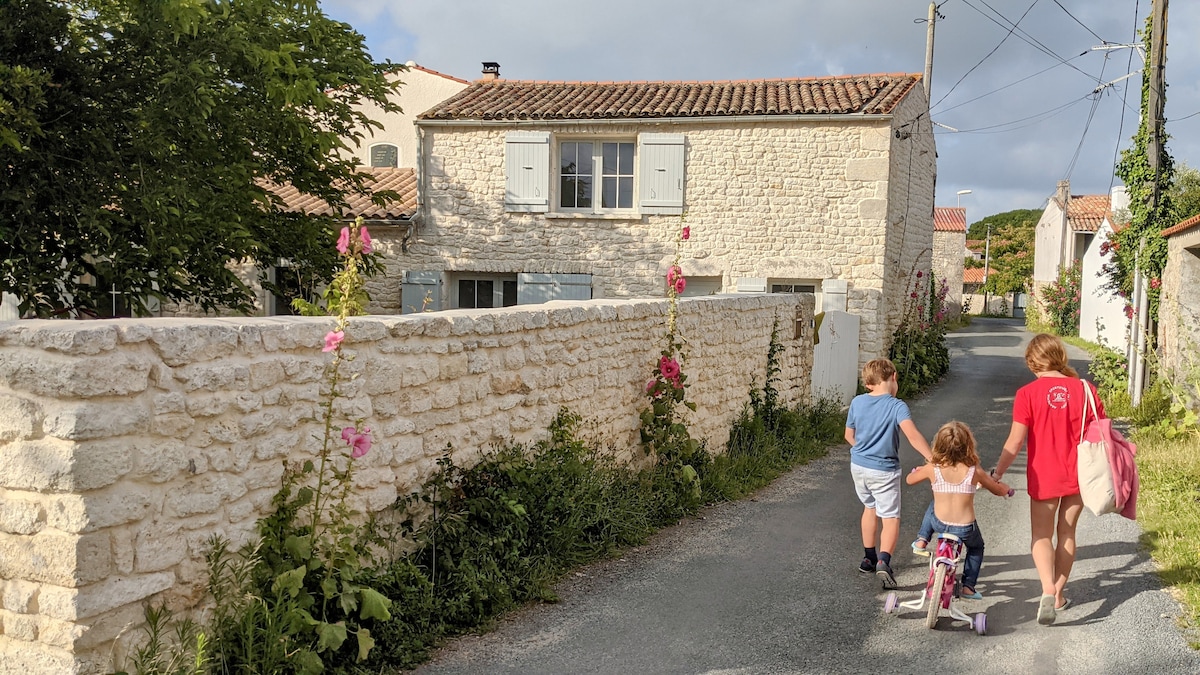 A quiet street lined with traditional stone houses is depicted. A family can be seen walking together, with a child riding a tricycle. Bright flowers grow along the roadside, adding color to the serene setting.