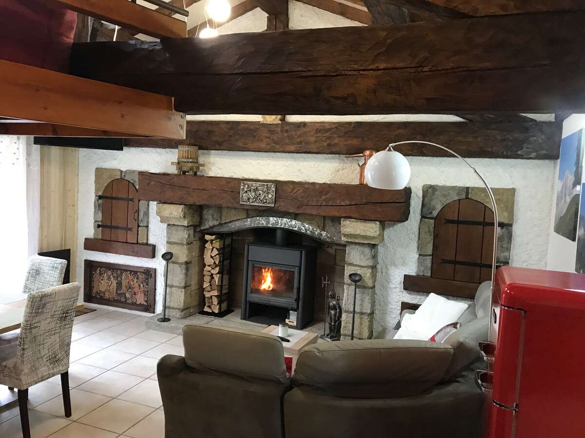 A cozy living space features a stone fireplace with a lit fire, surrounded by wooden beams and rustic decor. A comfortable seating area with pale cushions is visible, along with a dining table and light-colored chairs. Natural light is filtered through nearby windows.
