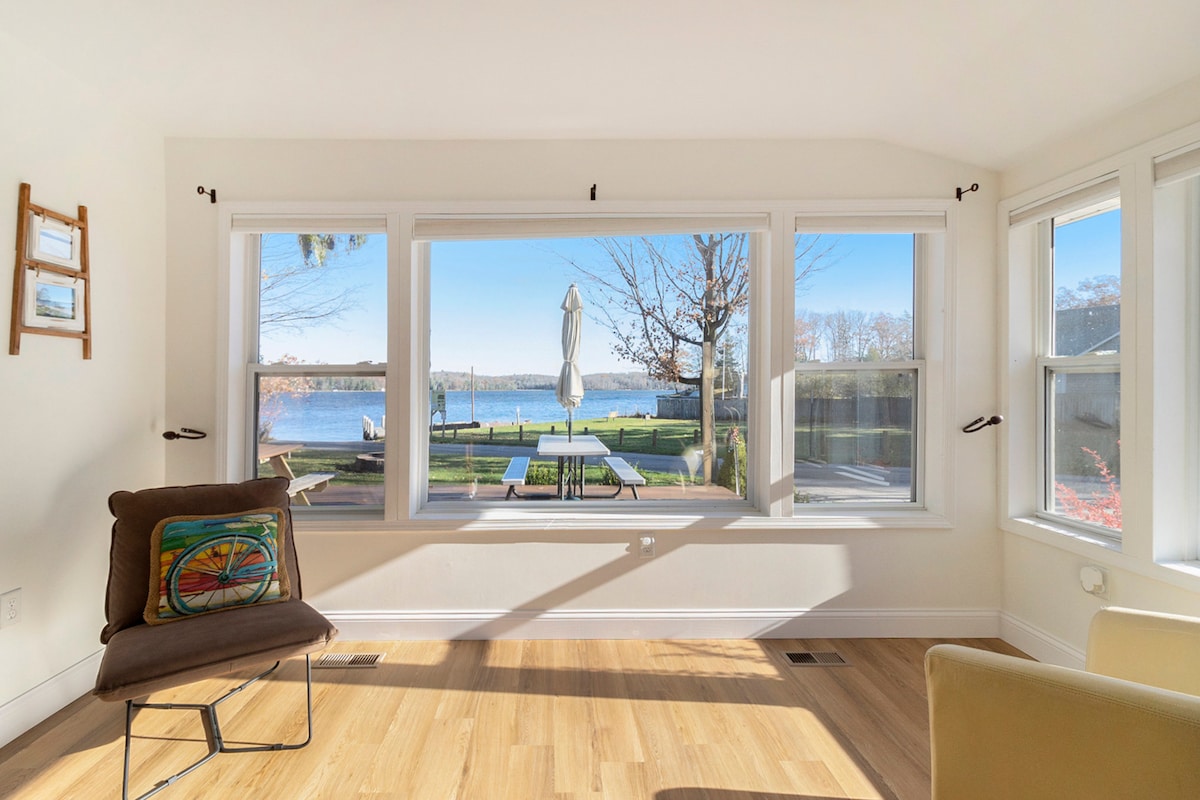 A bright living area features large windows offering unobstructed views of Hamlin Lake. A small chair with a colorful design adds a cozy touch. Light wooden flooring enhances the open feel, creating a welcoming space for relaxation.