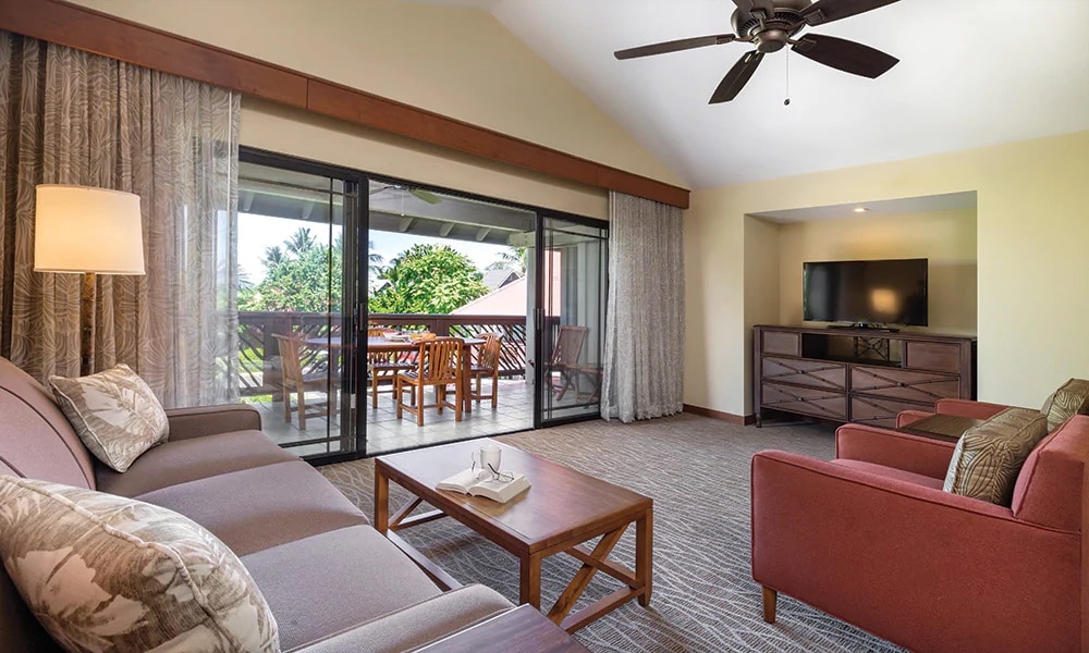 A comfortable living area features a sofa and an armchair arranged around a wooden coffee table. Natural light floods in through glass doors that open to a balcony with outdoor seating. A ceiling fan and TV are also visible, providing a cozy and inviting space.